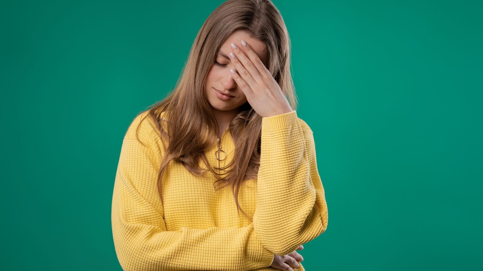 A woman in a yellow sweater stands before a green background, covering her face with her hand and looking stressed.