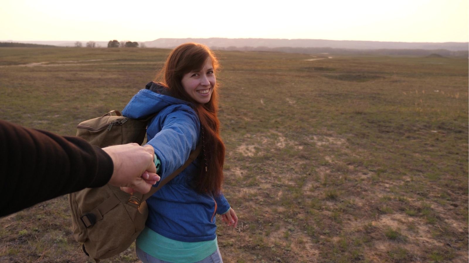 A smiling woman with a backpack holds someone's hand as they walk through a grassy landscape at sunset.