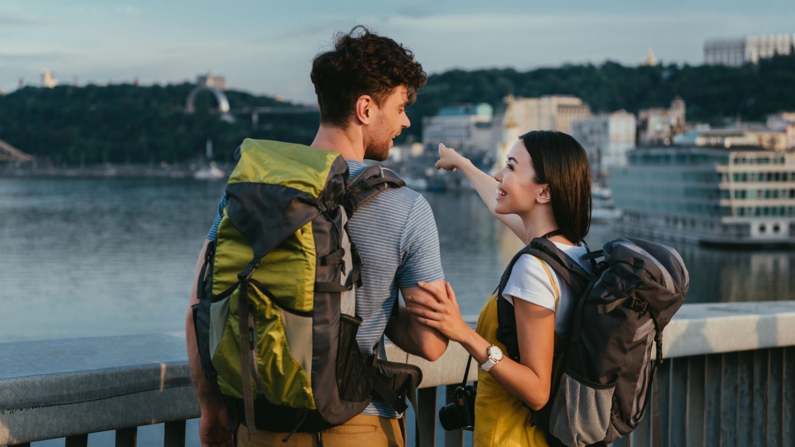 Two backpackers stand on a bridge over a river, one pointing toward the city skyline as the other looks that way.