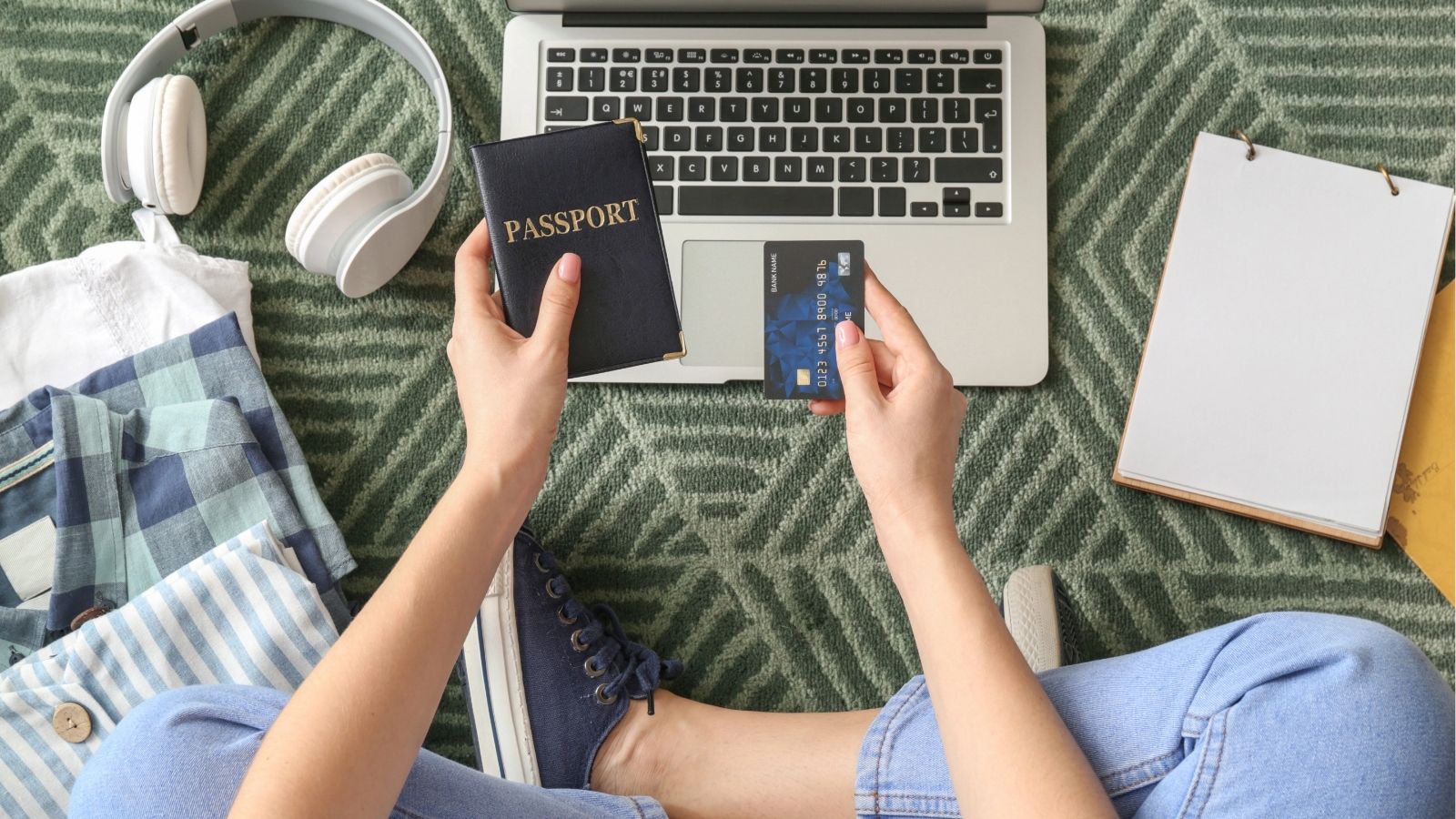 Person on a green carpet with a laptop, passport, and credit card amid headphones, clothes, and a notepad.