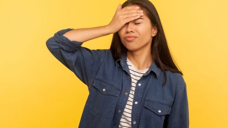 A woman with eyes closed and hand on her forehead stands before a yellow background, looking frustrated.