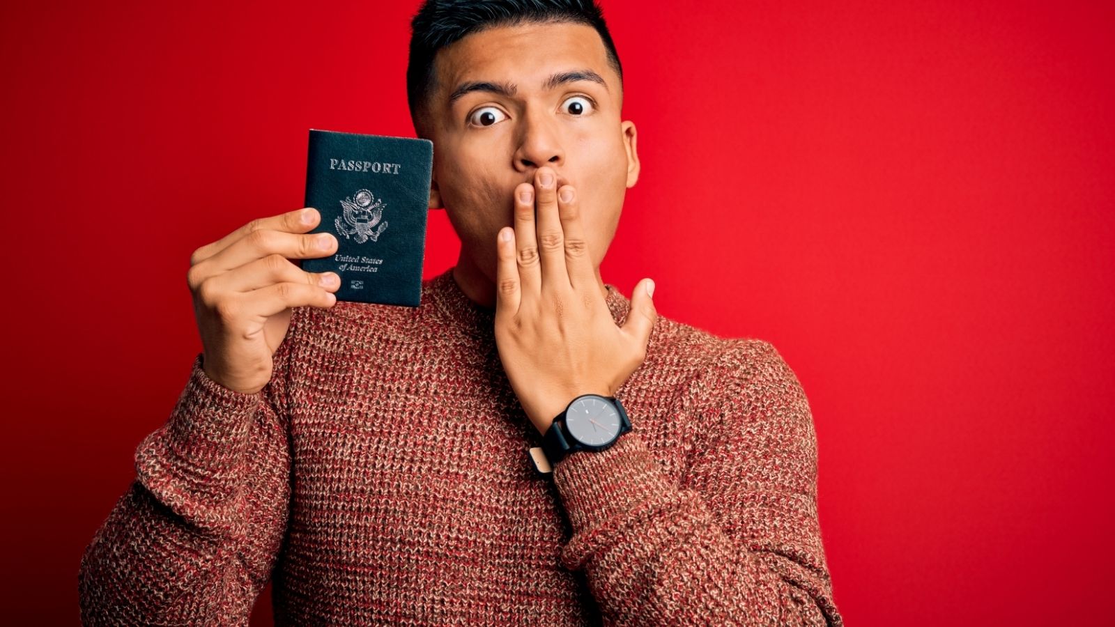 A person in a brown sweater covers their mouth with one hand and holds up a U.S. passport against a red background.