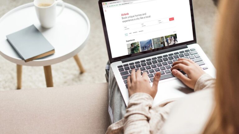 A person sits with a laptop on their lap viewing Airbnb, next to a round side table holding a notebook and cup.