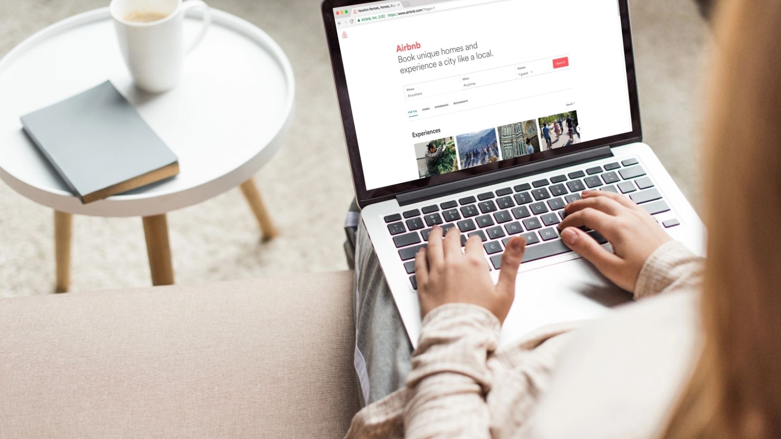 A person sits with a laptop on their lap viewing Airbnb, next to a round side table holding a notebook and cup.