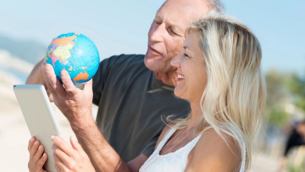 An older man and woman smile outdoors as he holds a globe and she holds a tablet, appearing to discuss travel plans.