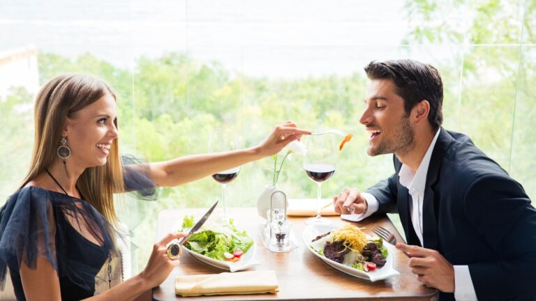 A woman feeds a man at a restaurant table with wine and meals, large windows showing greenery in the background.