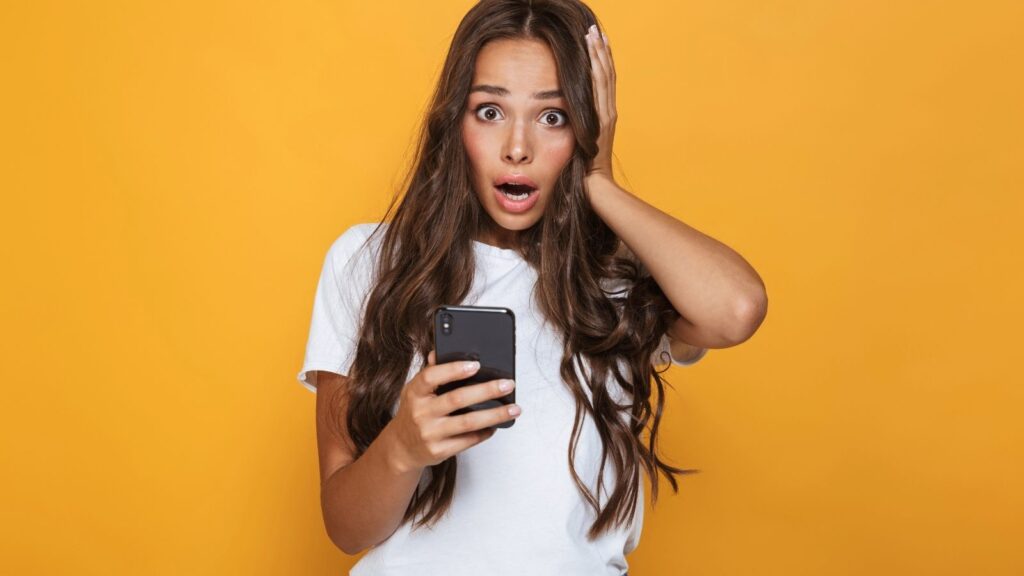 Surprised woman with long brown hair holds a smartphone and touches her face, standing before a yellow background.