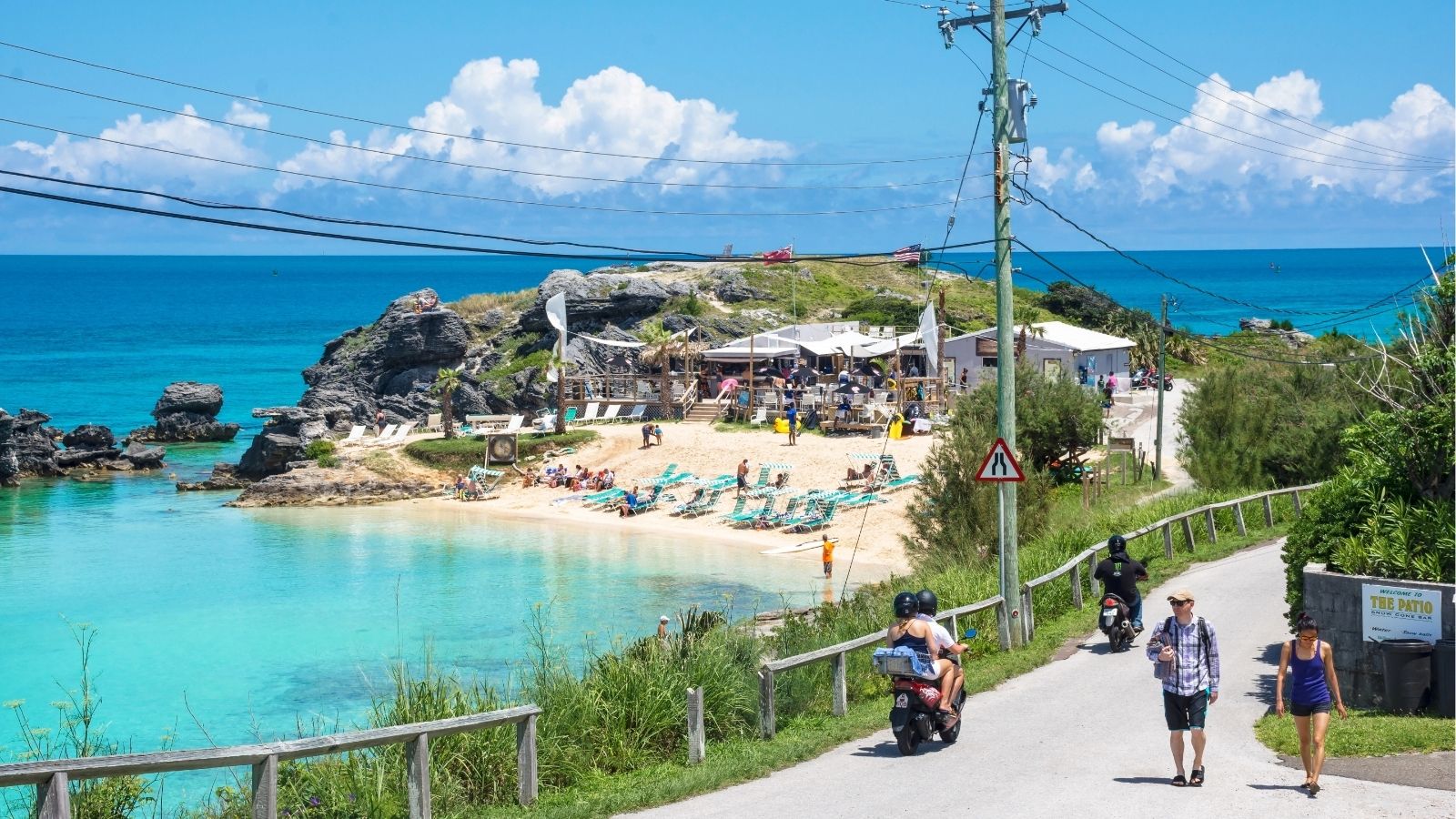 A small, turquoise beach with sunbathers is bordered by cliffs and buildings, with people walking and riding scooters in front.