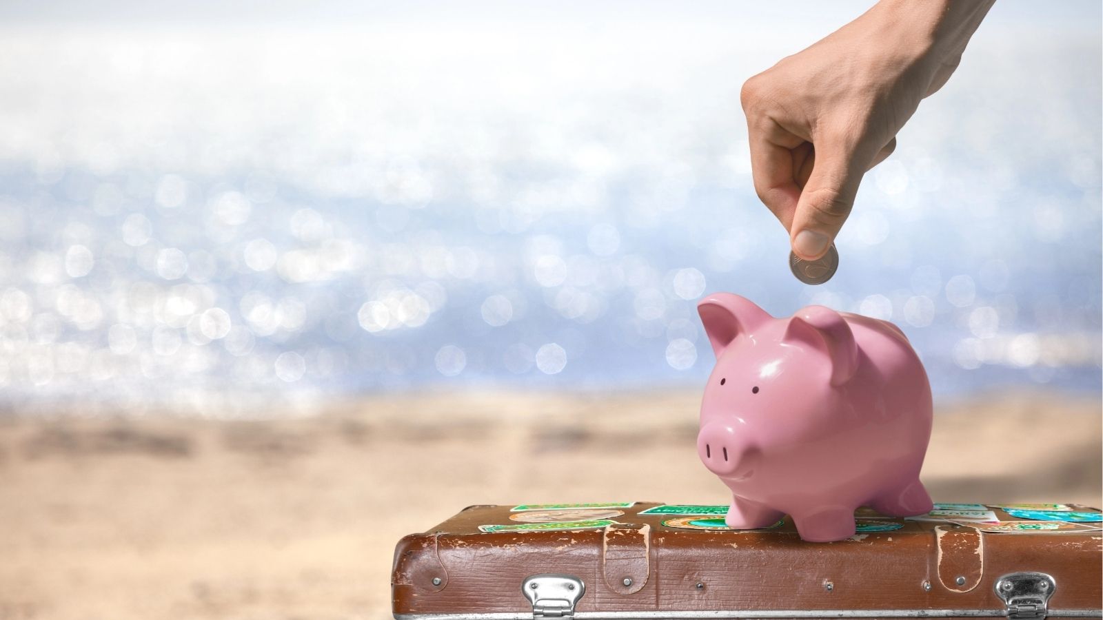 A hand puts a coin in a pink piggy bank on a suitcase, with sandy beach and ocean in the background.