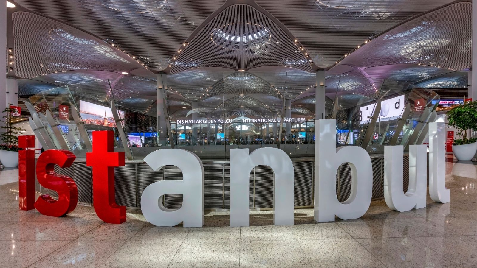 Large red and white "Istanbul" sign in a modern airport terminal, with an "International Departures" sign in the background.