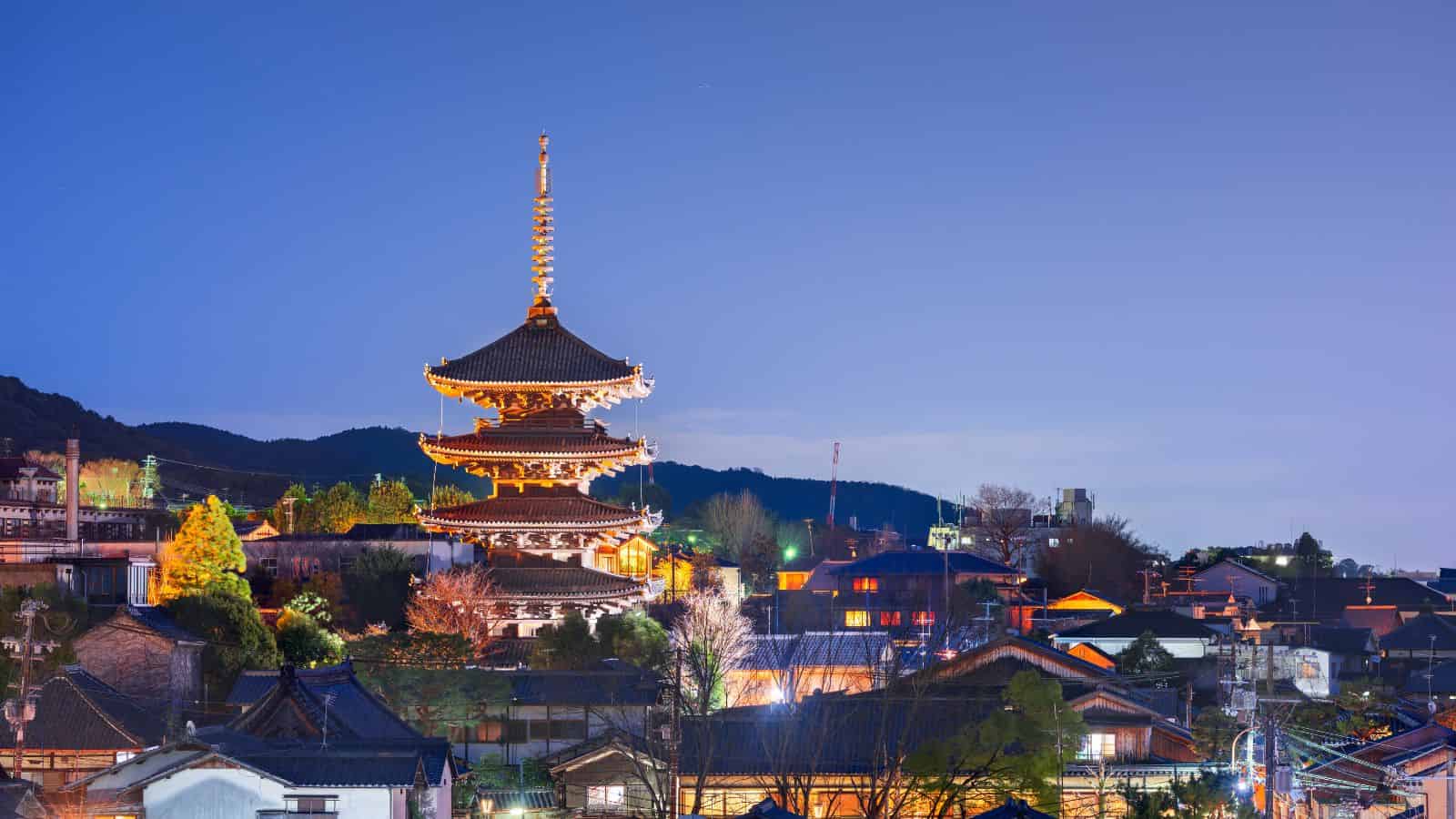 An illuminated five-story pagoda stands at dusk among rooftops and trees, with mountains in the background.