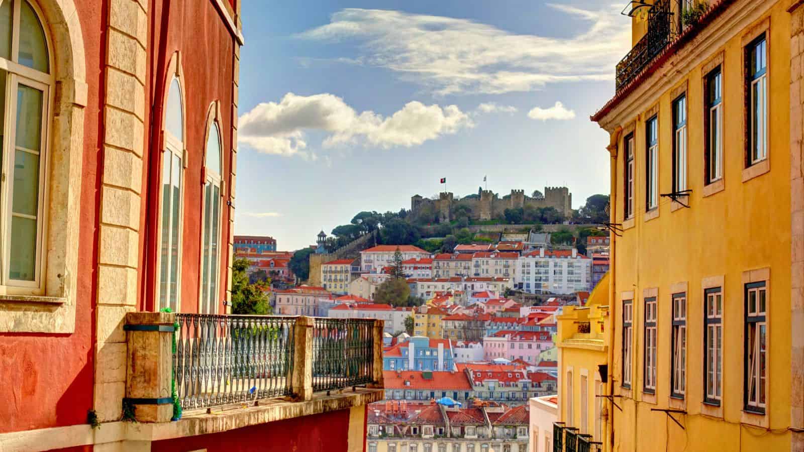Colorful buildings line a narrow street overlooking a hillside castle and cityscape beneath a partly cloudy sky.