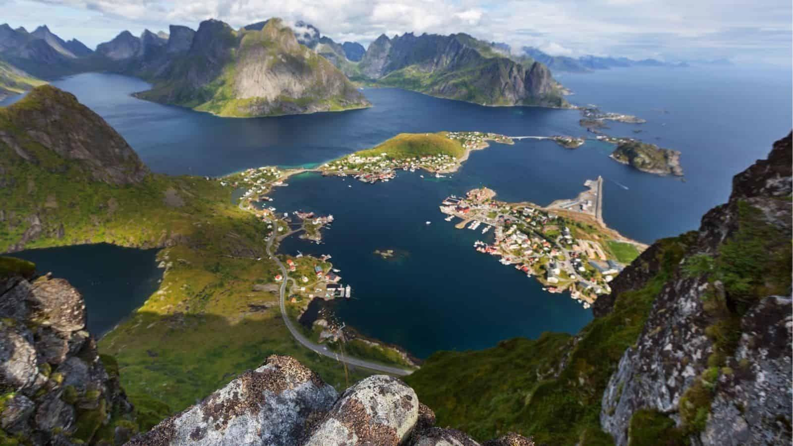Aerial view of a coastal village with scattered houses, roads, and a harbor amid mountains and fjords under partly cloudy skies.