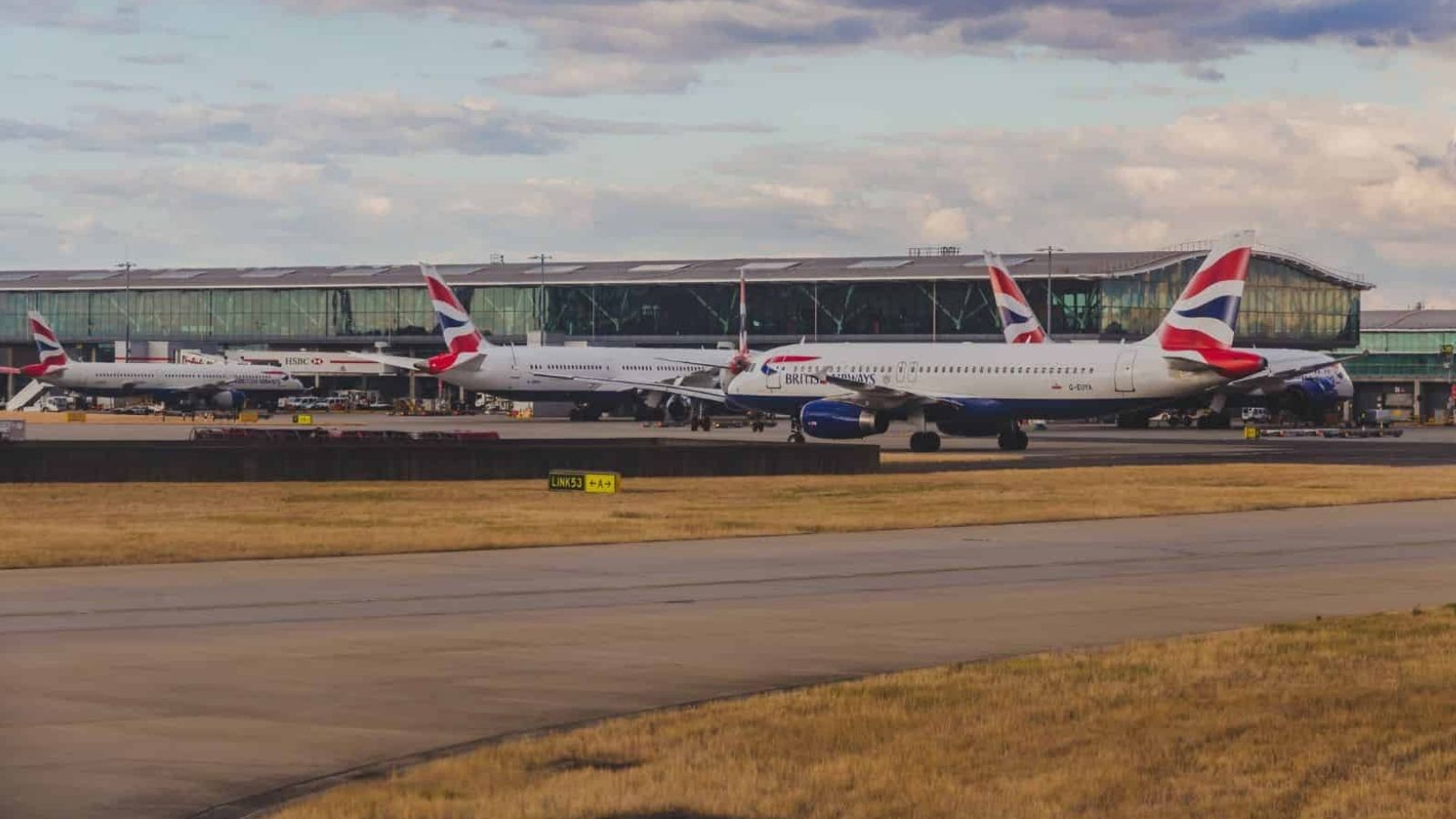 British Airways planes are parked at airport gates by a glass terminal beneath a partly cloudy sky.