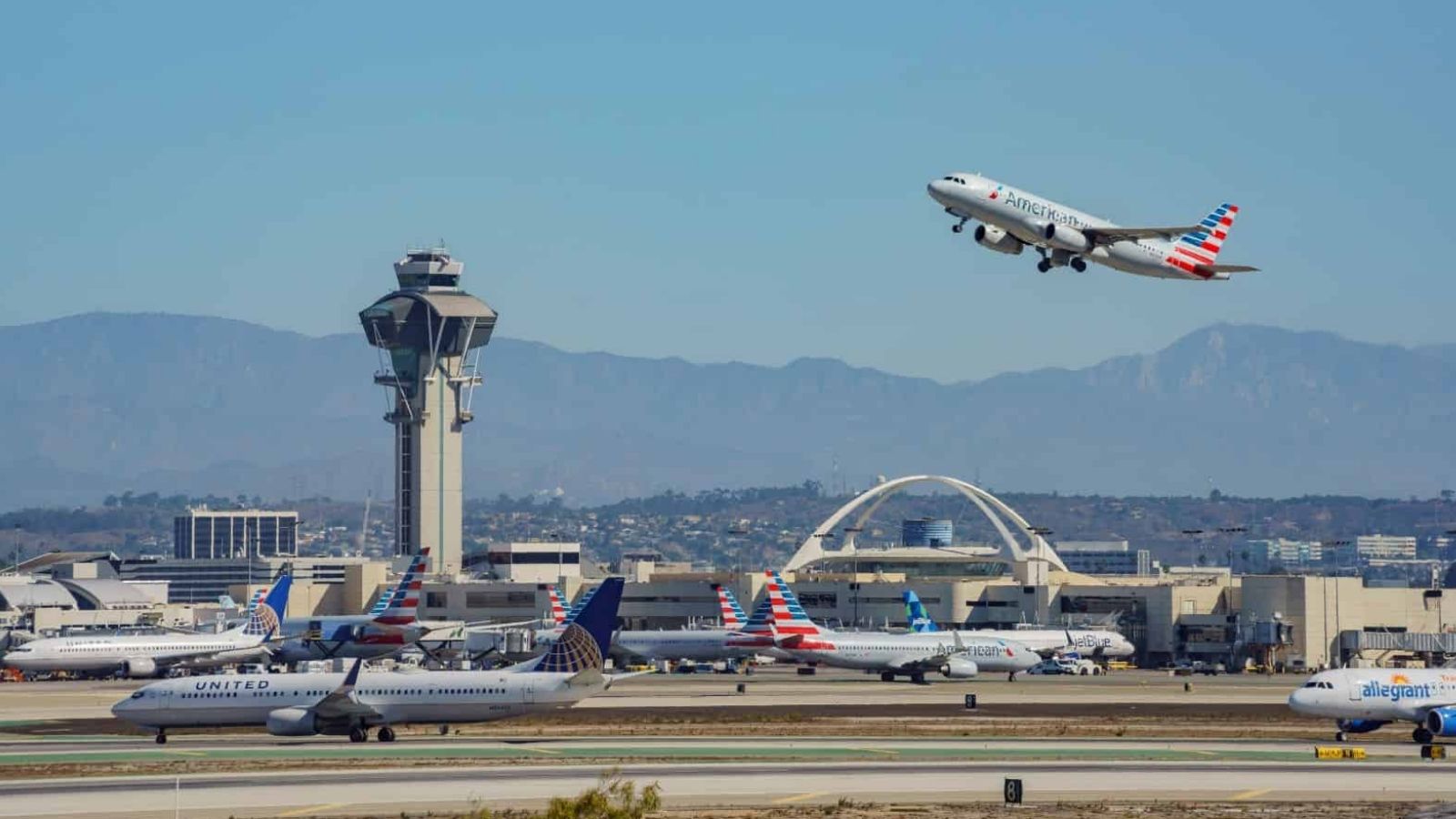 Commercial airplanes are on a busy airport runway, including one taking off, with the control tower and terminals in the background.