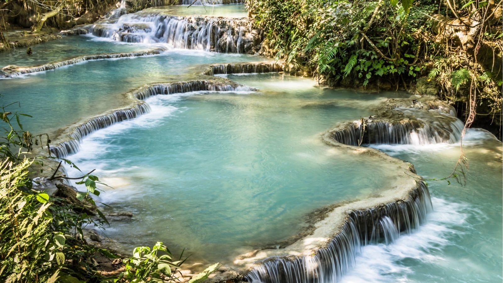 Turquoise terraced pools cascade over limestone, bordered by lush green vegetation.