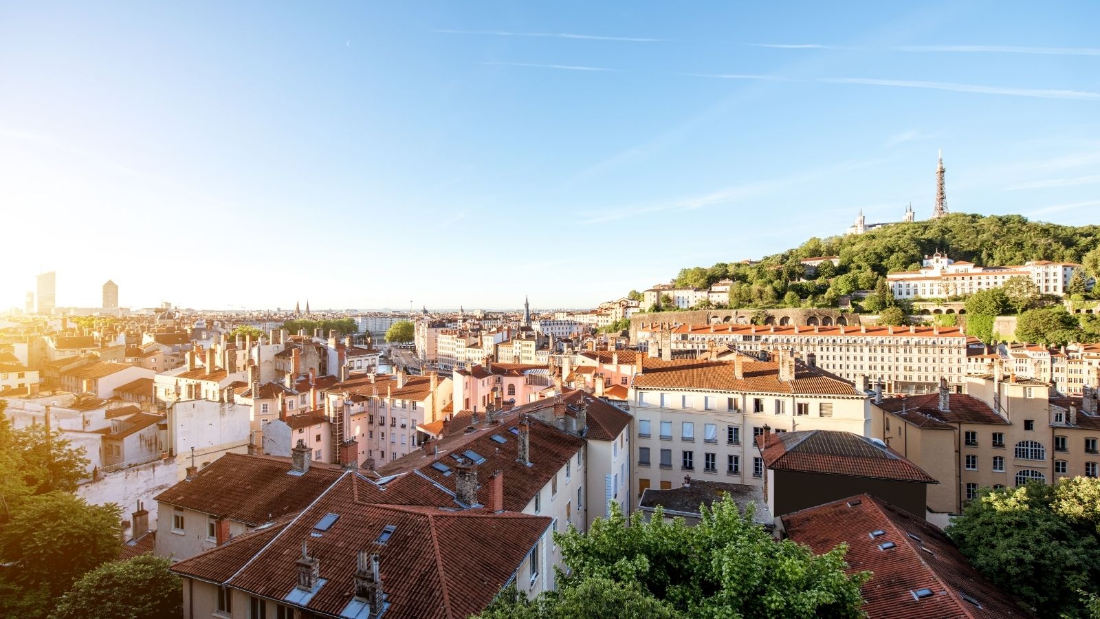City panorama with red-tiled roofs, dense buildings, and a hilltop basilica amid greenery beneath a clear sky.