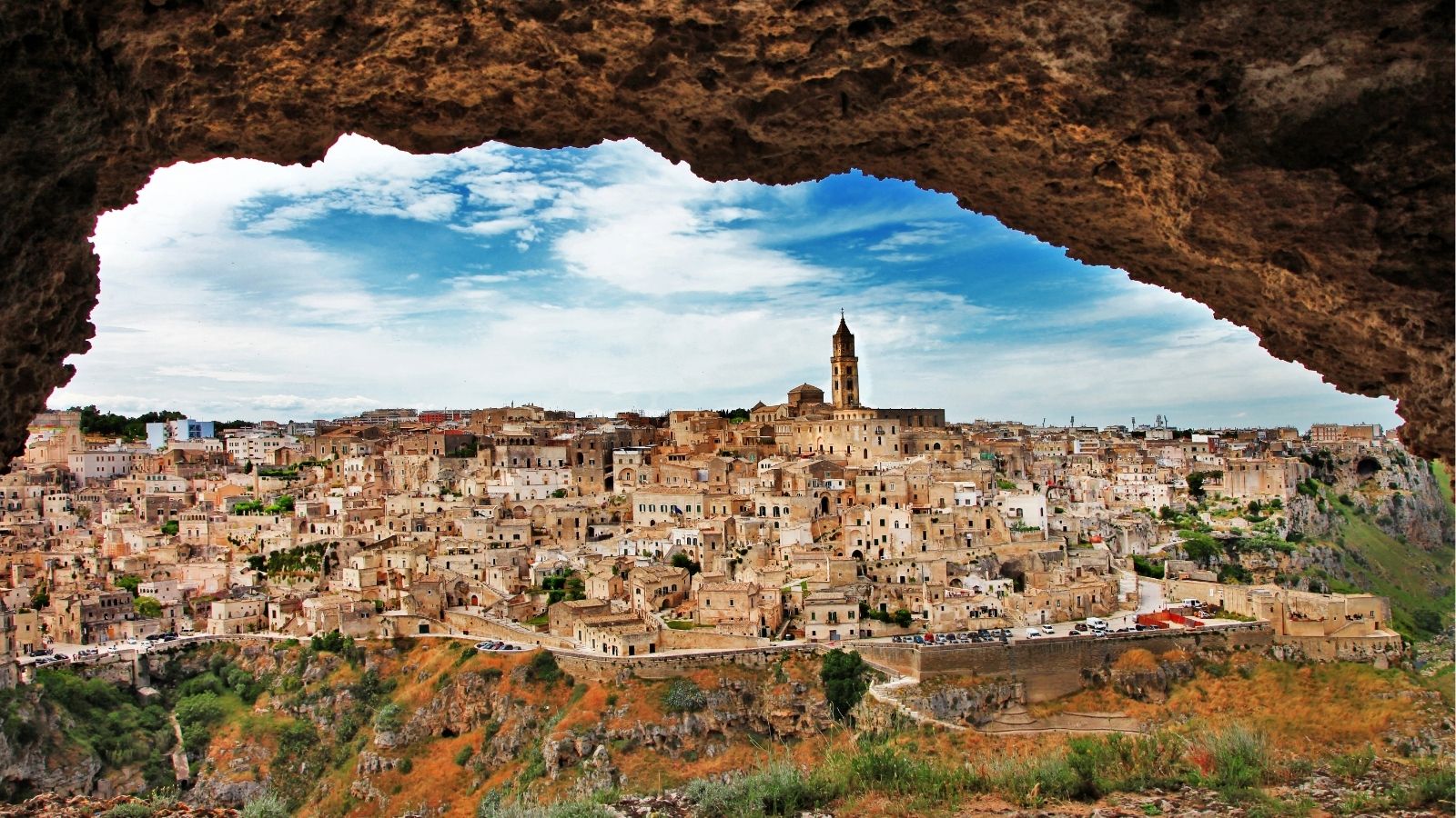 Matera, Italy’s stone buildings and church seen through a rocky cave opening beneath a partly cloudy sky.