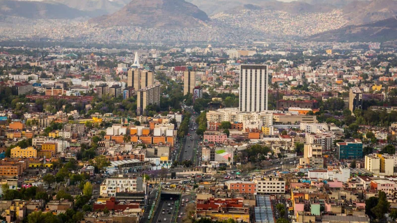 Aerial view of a crowded city with skyscrapers, busy roads, and distant mountains beneath a hazy sky.