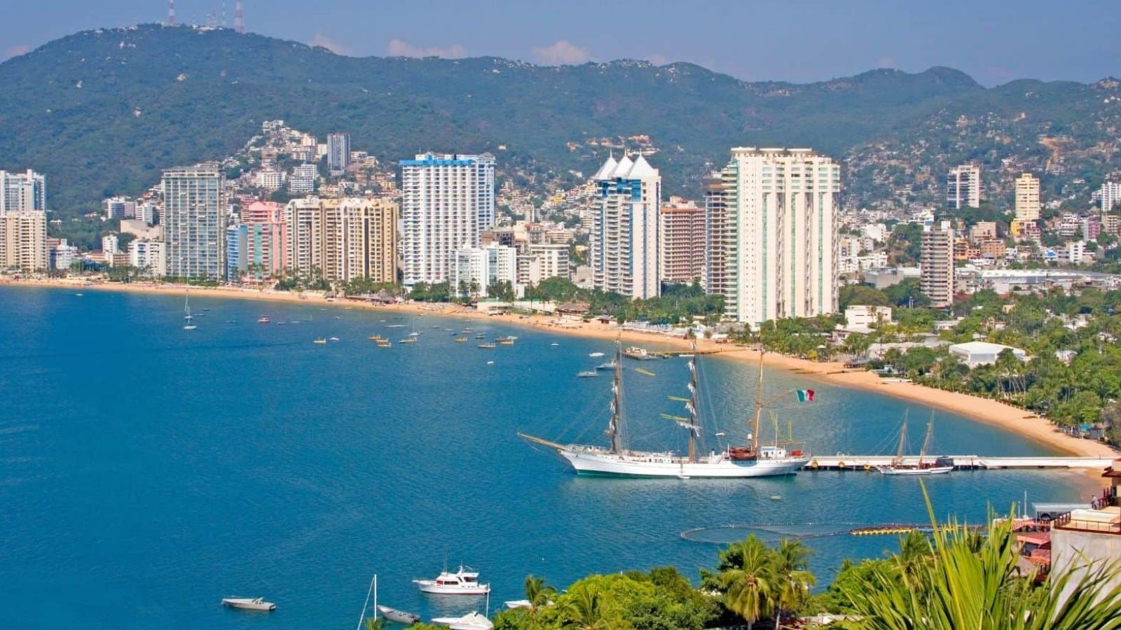 Tall buildings line a curved sandy beach with boats on the water and green hills in the background.