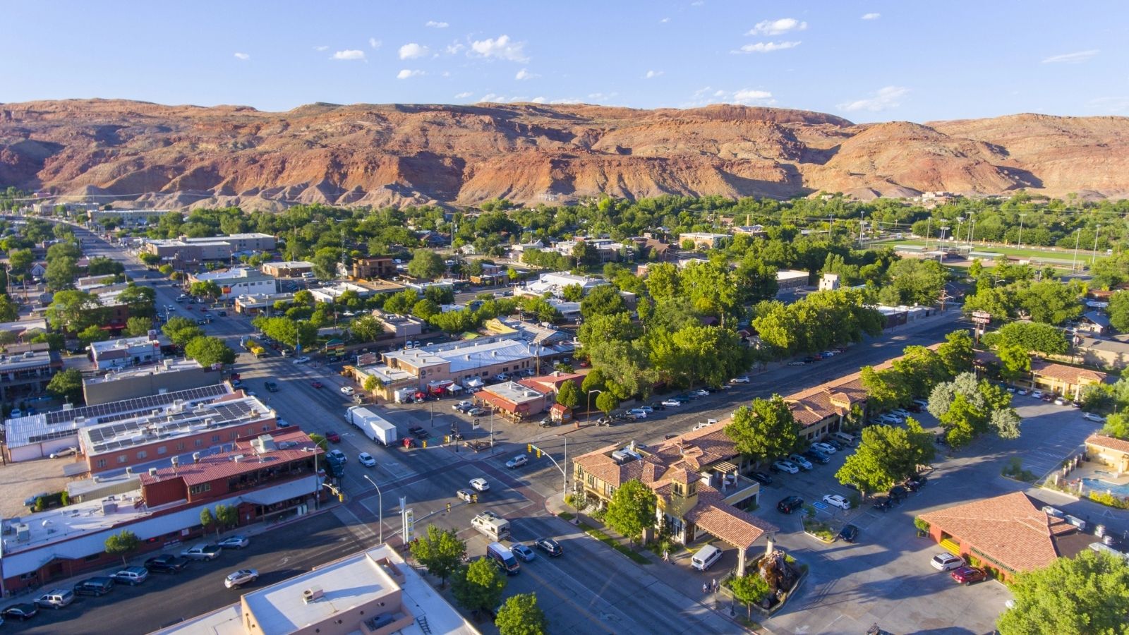 Aerial shot of a small town with tree-lined streets and buildings, bordered by red hills under a clear blue sky.