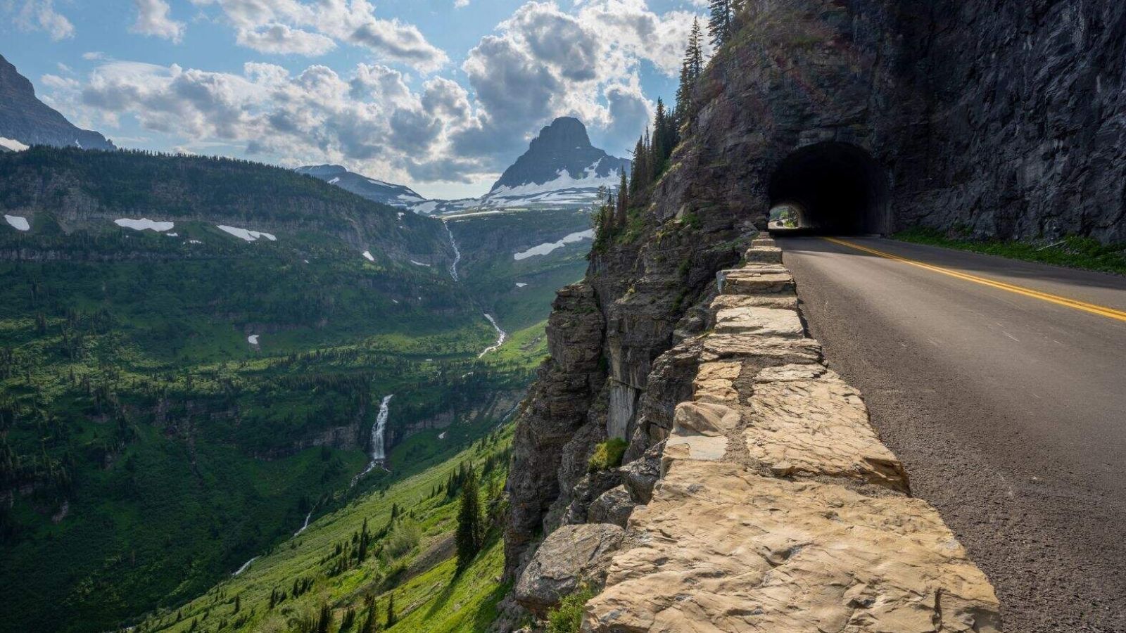 A mountain road enters a cliffside tunnel, overlooking a green valley with waterfalls and snowy peaks beneath a partly cloudy sky.