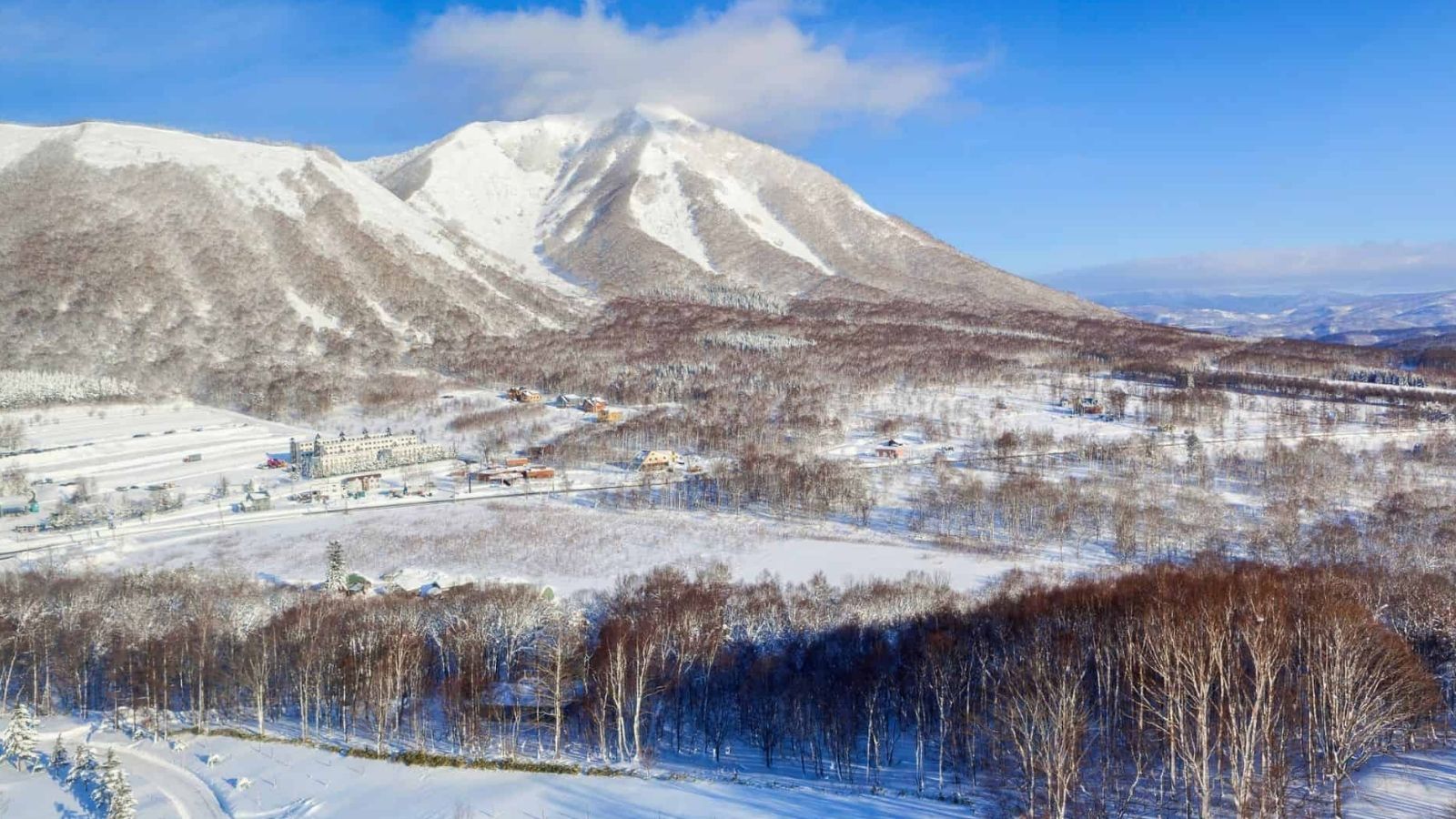 Snow blankets a landscape of scattered buildings and bare trees, set before distant mountains beneath a clear blue sky.