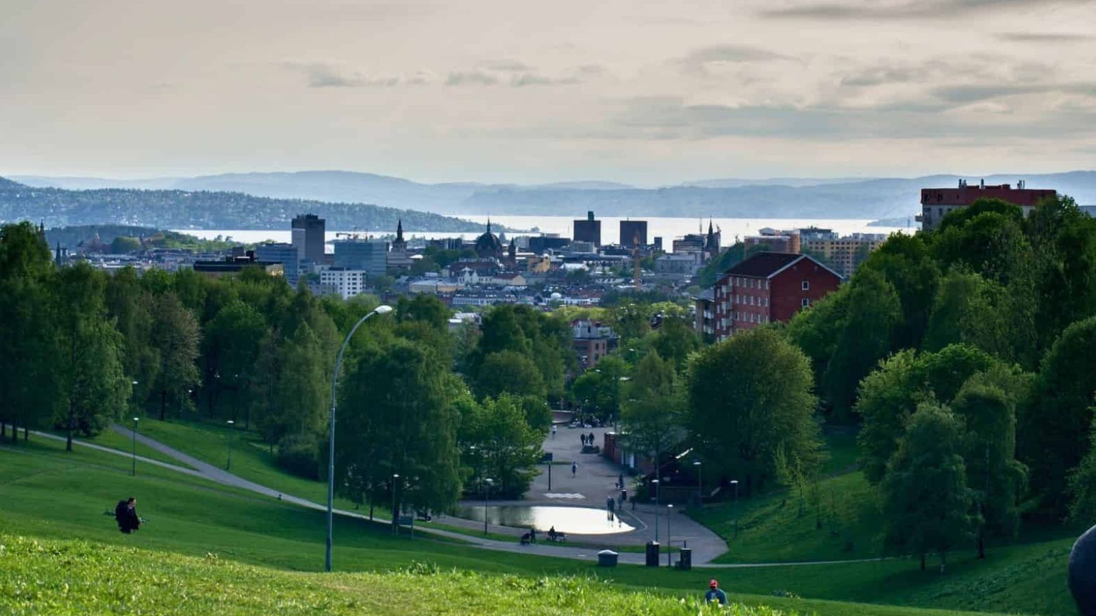 Distant city buildings rise beyond green trees and a park in the foreground beneath a cloudy sky.