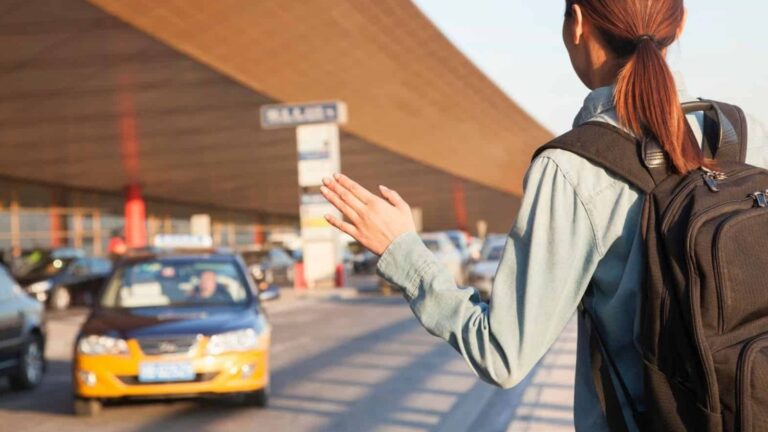 A backpacked person hails a taxi outside an airport terminal, with cars and signs in the background.