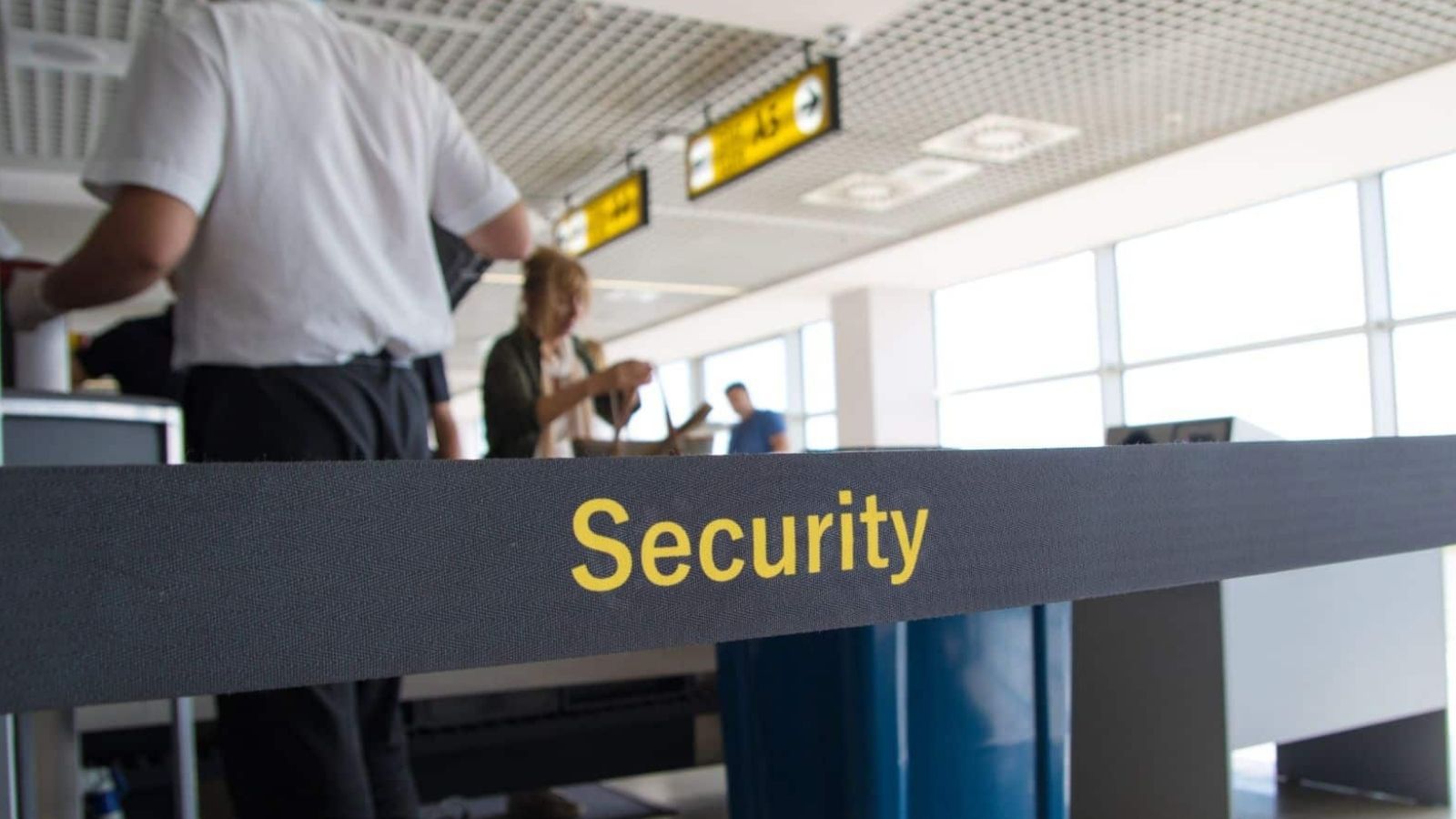 A black "Security" belt in yellow letters marks an airport checkpoint, with people and signs visible behind it.
