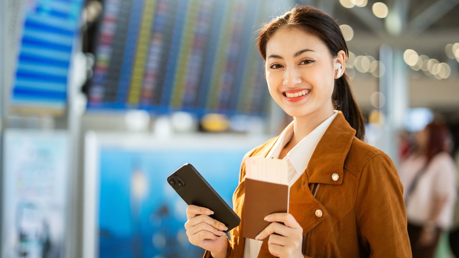 Smiling woman at airport holds smartphone and passport with boarding pass; flight info boards appear in the background.