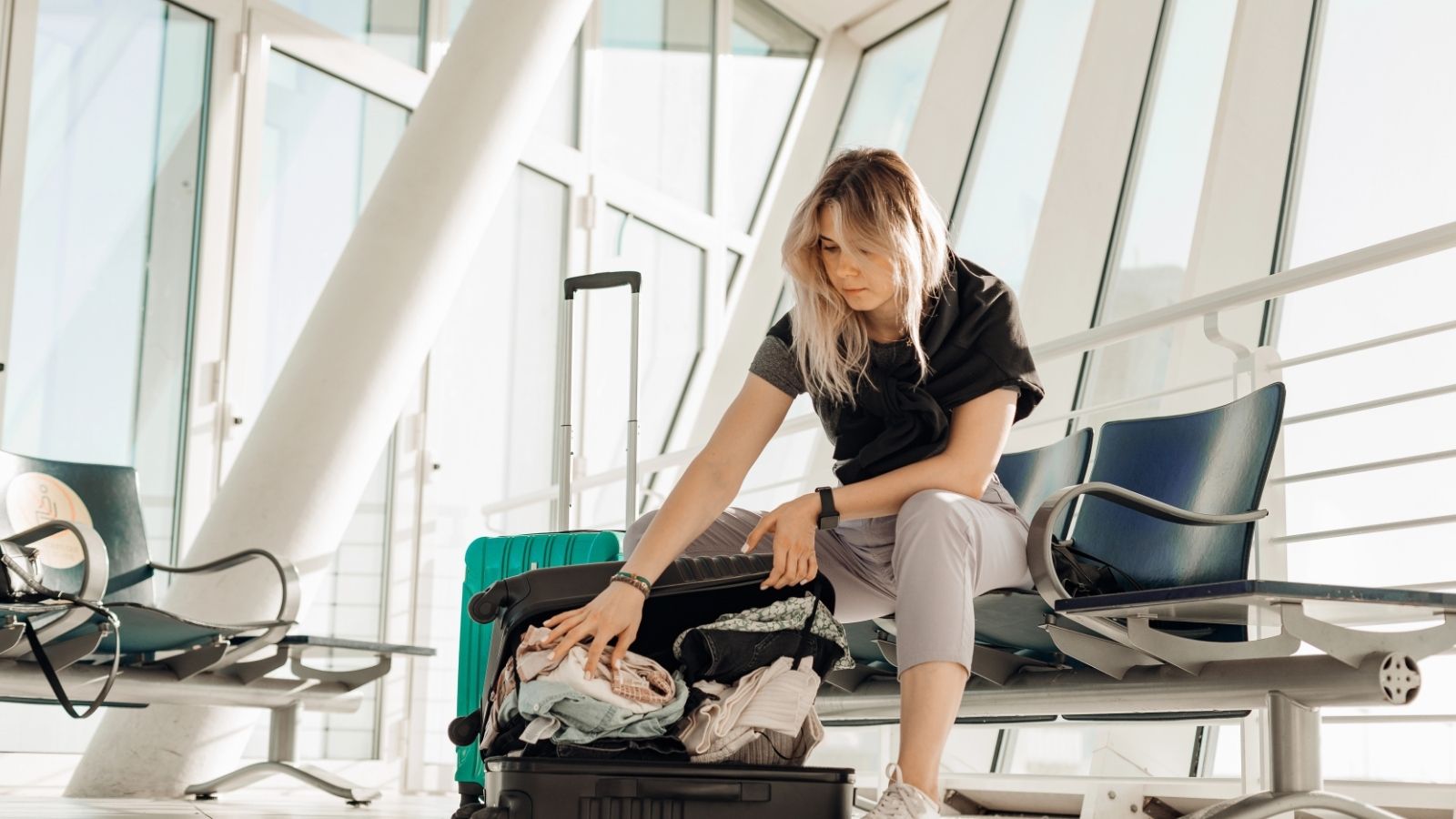 A woman on an airport bench organizes clothes in an open suitcase beside a green rolling suitcase.