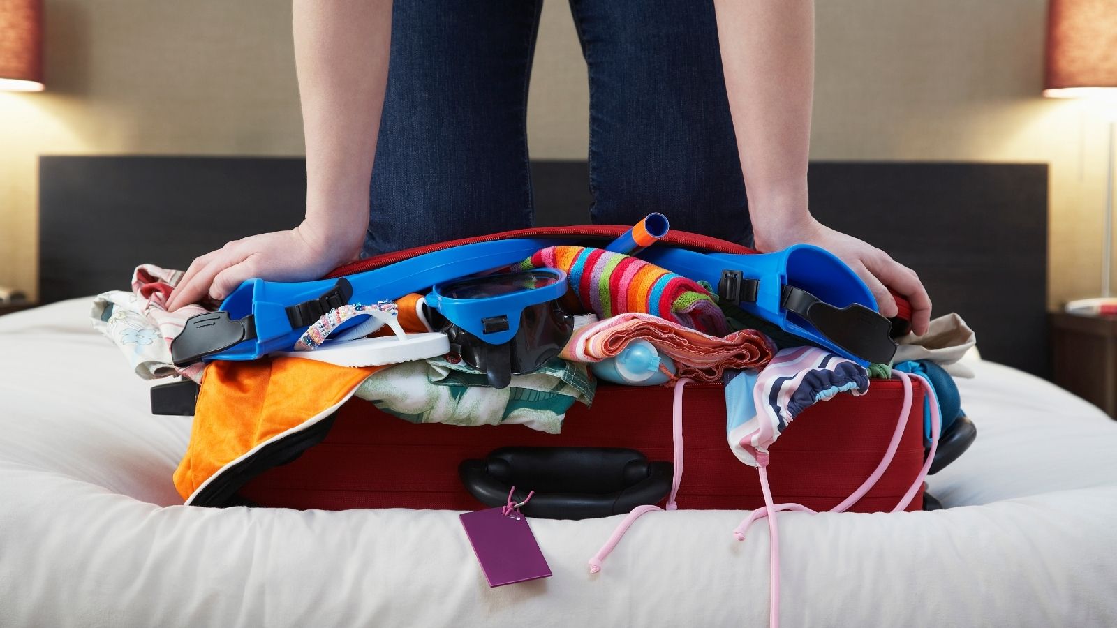 Person pressing down an overstuffed suitcase on a bed, with colorful clothes and swim gear spilling out.