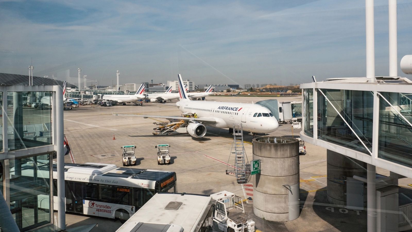 An Air France plane is at an airport gate with boarding bridges, service vehicles, and other aircraft in the background.