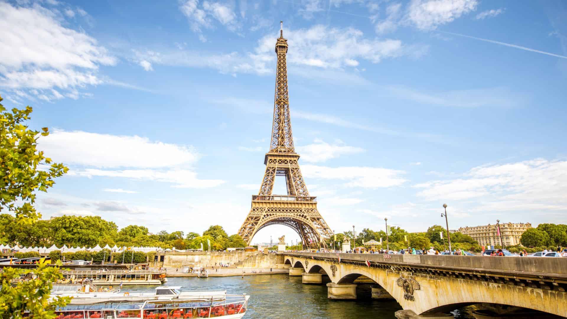 The Eiffel Tower rises by the Seine in Paris, with a bridge, trees, and boats beneath a partly cloudy sky.