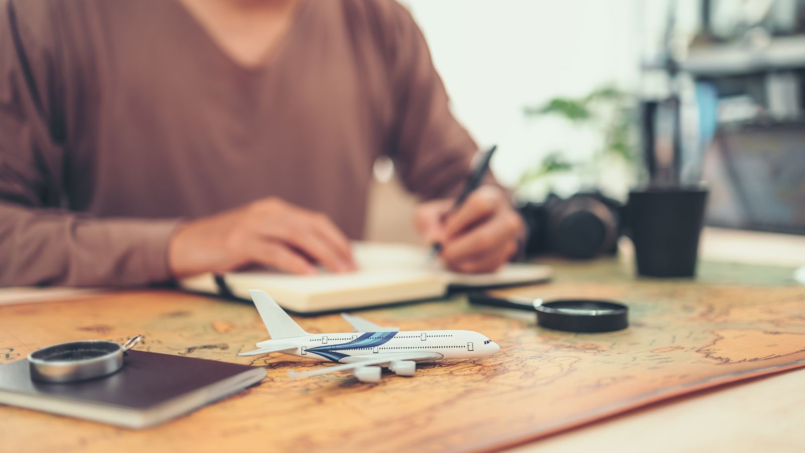 A person writes in a notebook with a toy airplane, passport, magnifying glass, and camera on a map in front.