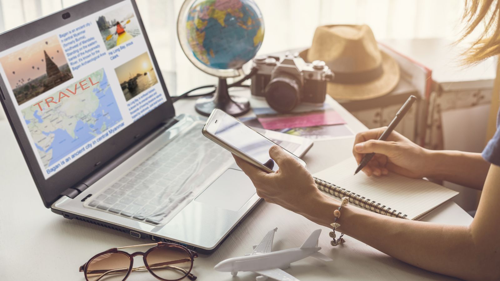 Person plans a trip at a desk with a laptop, notepad, phone, globe, camera, sunglasses, hat, and model airplane.