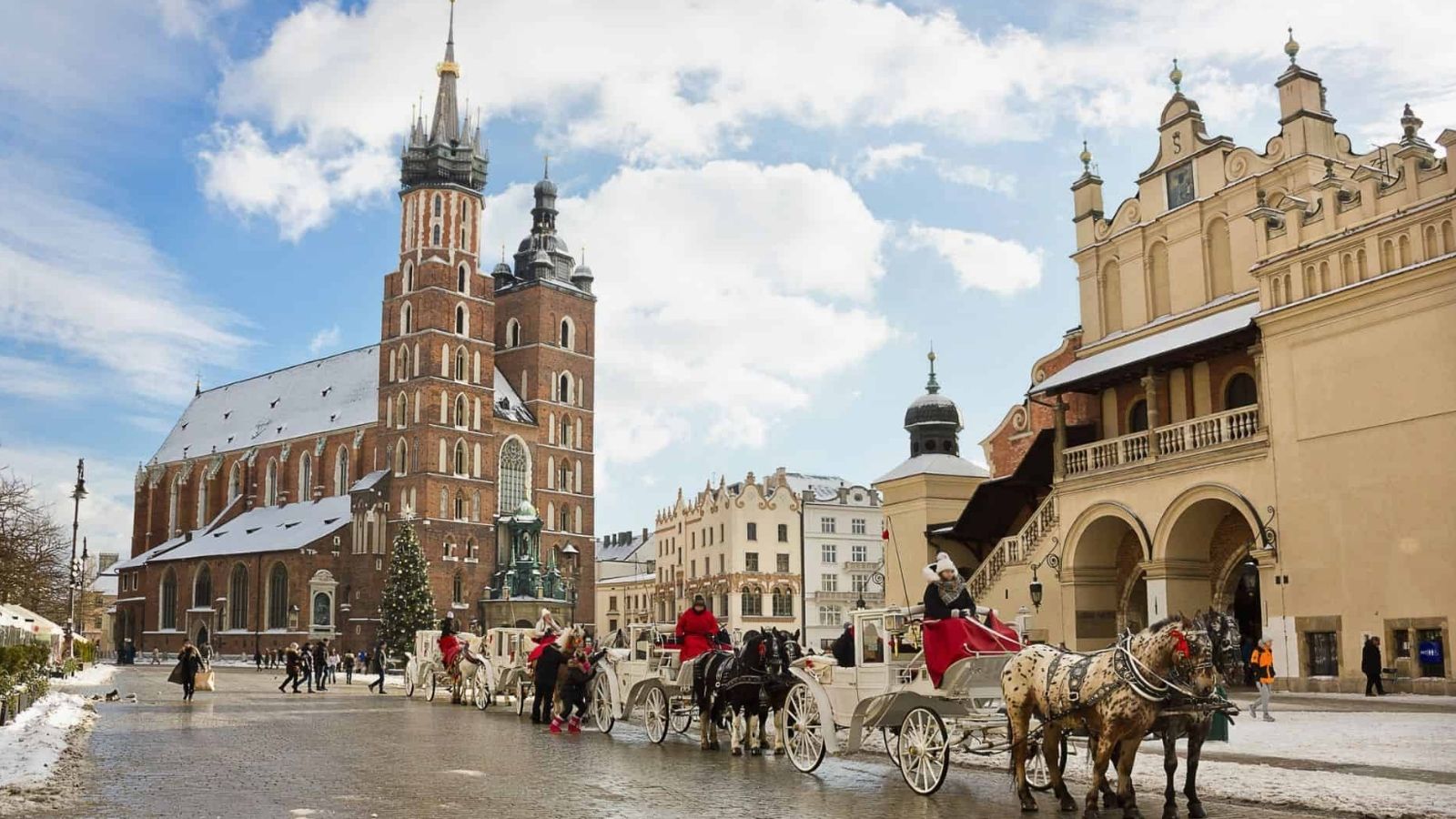 Horse-drawn carriages travel a cobblestone street past old buildings and a church in a historic city square on a winter day.