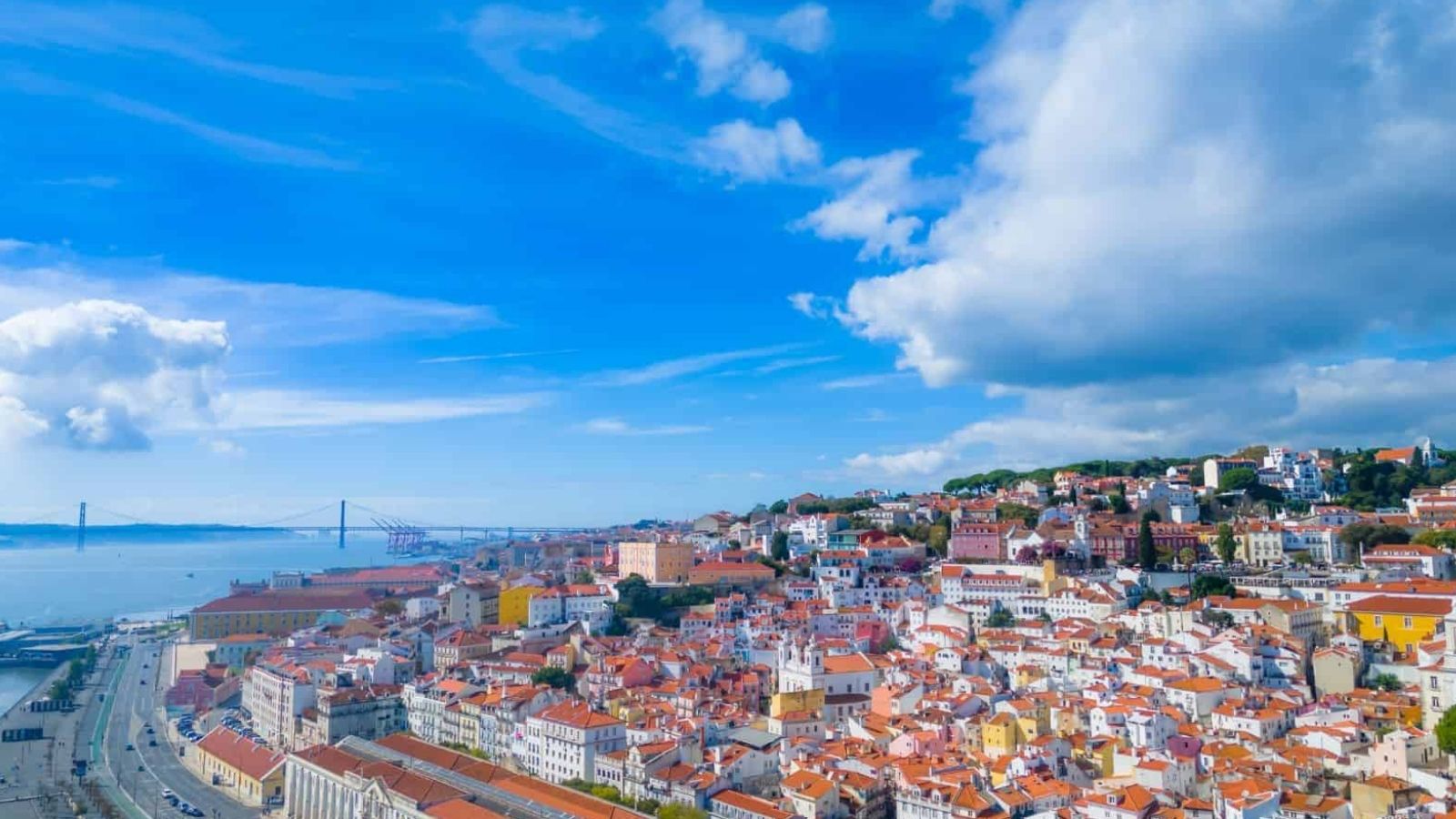 Aerial view of a coastal city with red roofs, a bridge in the background, and a partly cloudy blue sky.
