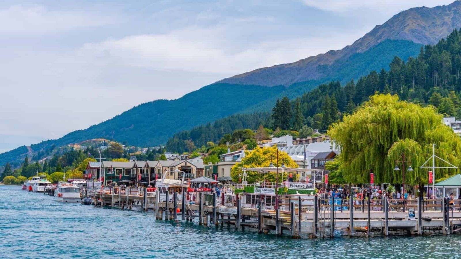 Waterfront with docks, boats, and buildings set before green trees, mountains, and a partly cloudy sky.