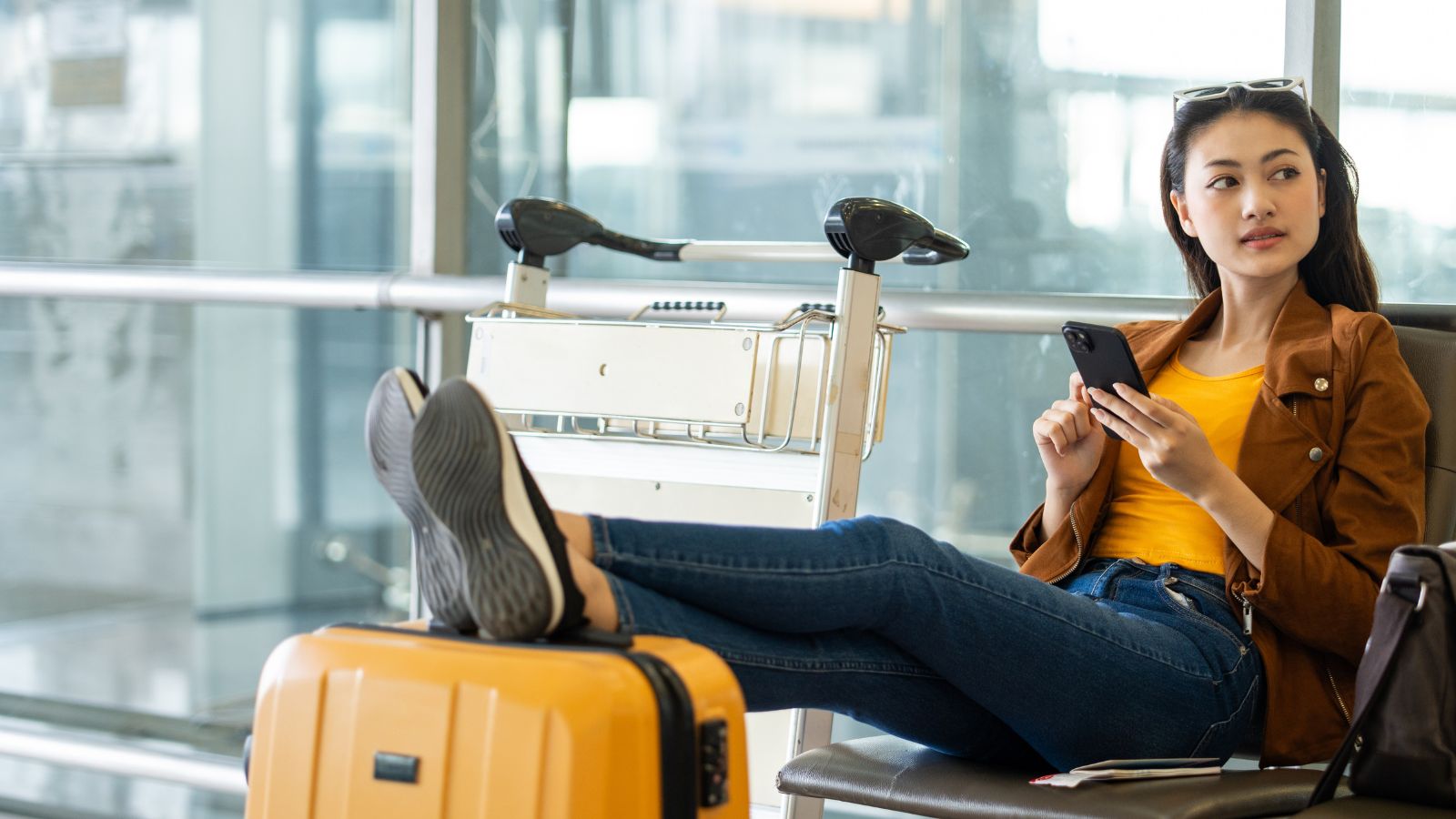 A woman relaxes in an airport lounge with her feet on a chair, holding a phone and a yellow suitcase in front of her.