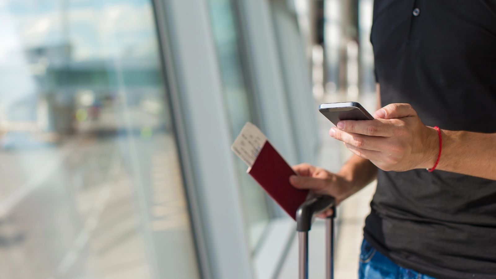 At an airport, a person holds a smartphone, passport with boarding pass, and grips a suitcase handle.