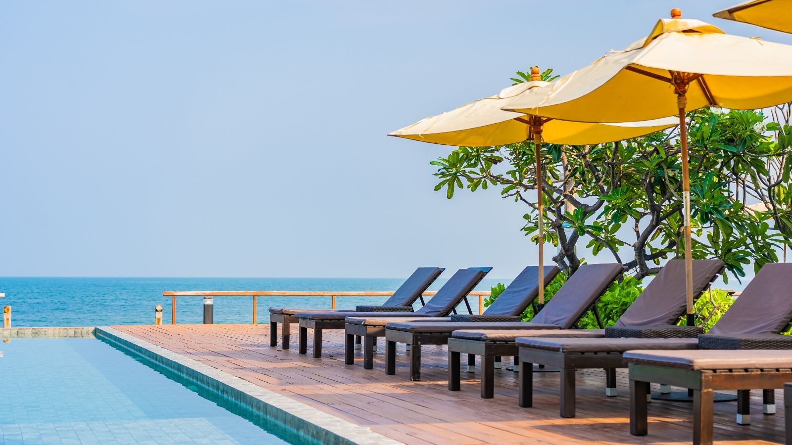 Empty lounge chairs with yellow umbrellas line a poolside wooden deck, facing the ocean beneath a clear sky.