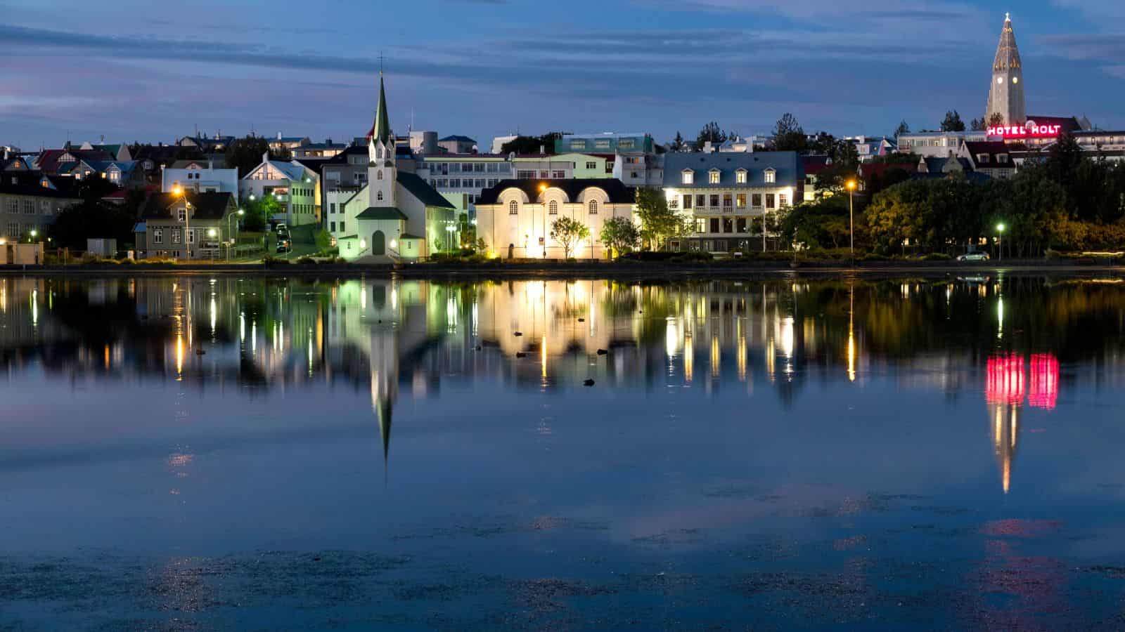 Dusk cityscape with buildings and a church reflected in calm water, illuminated windows mirrored on the surface.