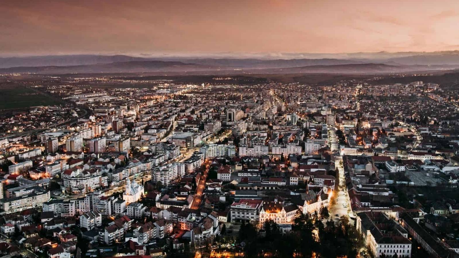 Aerial view of a city at dusk with illuminated buildings and streets beneath a cloudy sky.