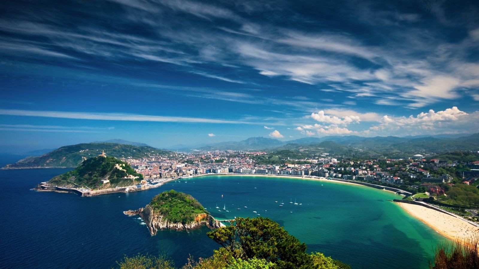 A coastal city features a crescent beach, green hills, and clear blue water beneath a partly cloudy sky.
