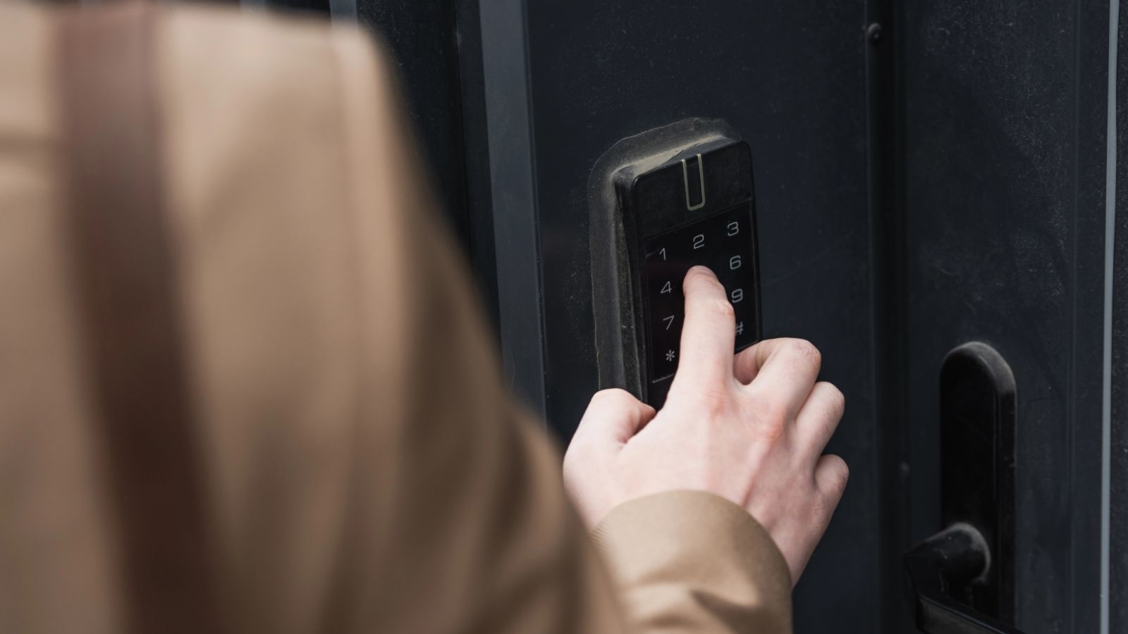 A person in a beige coat enters numbers on a black keypad lock attached to a dark door.