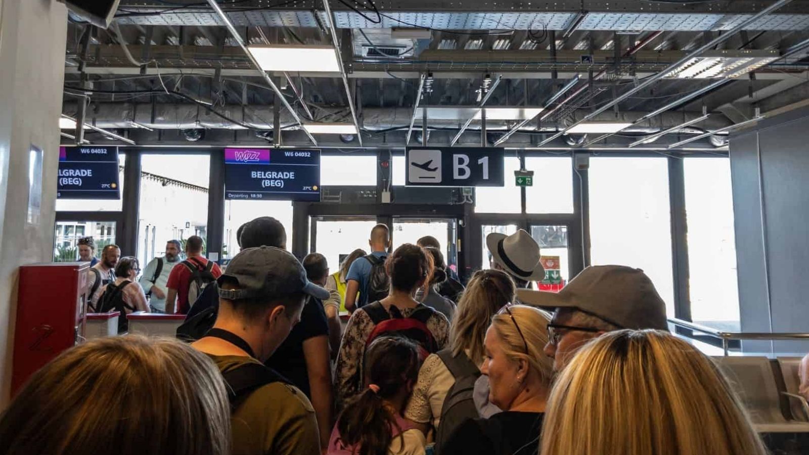 Travelers line up at airport gate B1 to board a flight to Belgrade, with signs and exposed ceiling structures overhead.