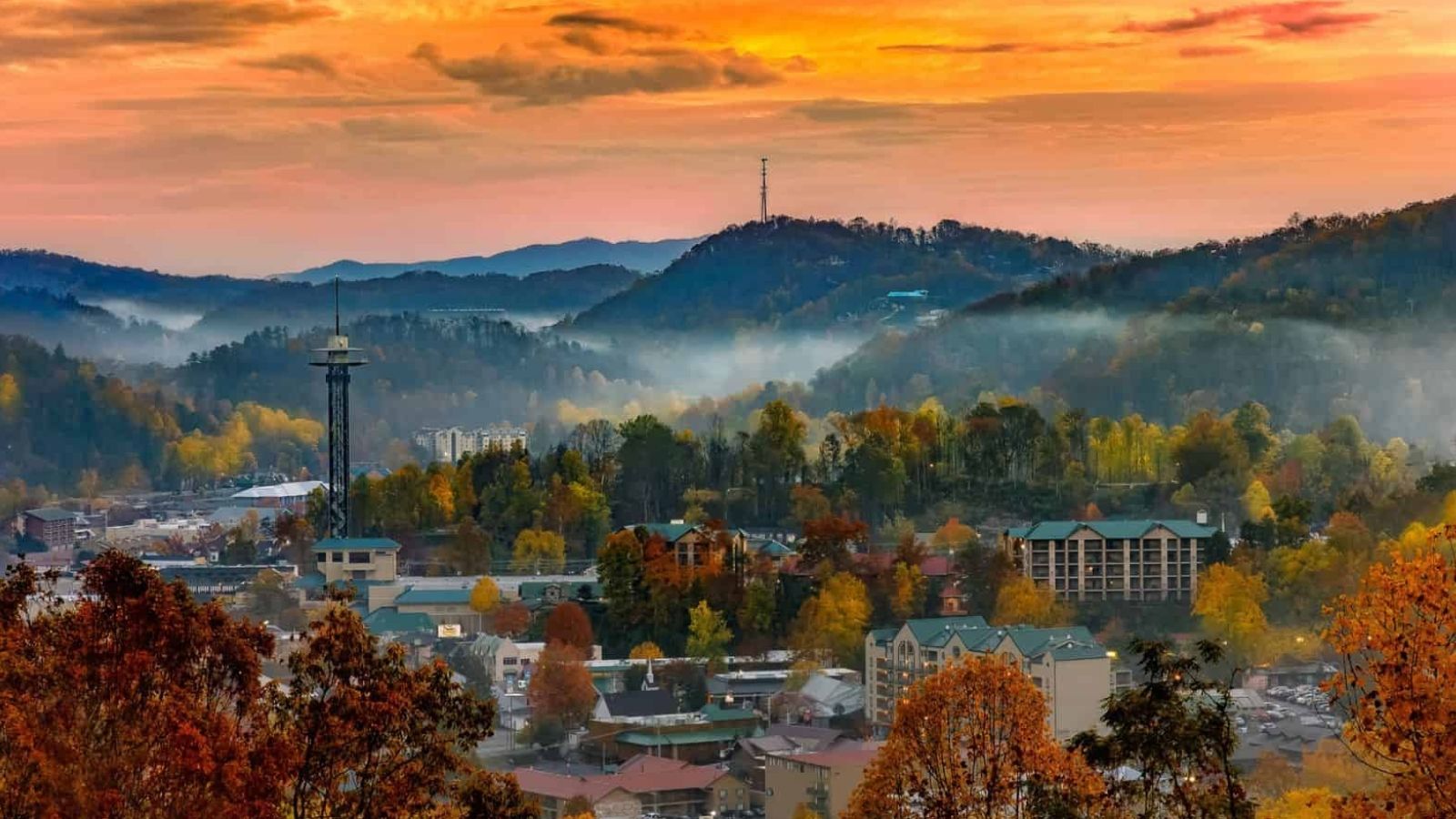 A small city nestled among autumn trees and hills, with mist and a colorful sunset sky in the background.