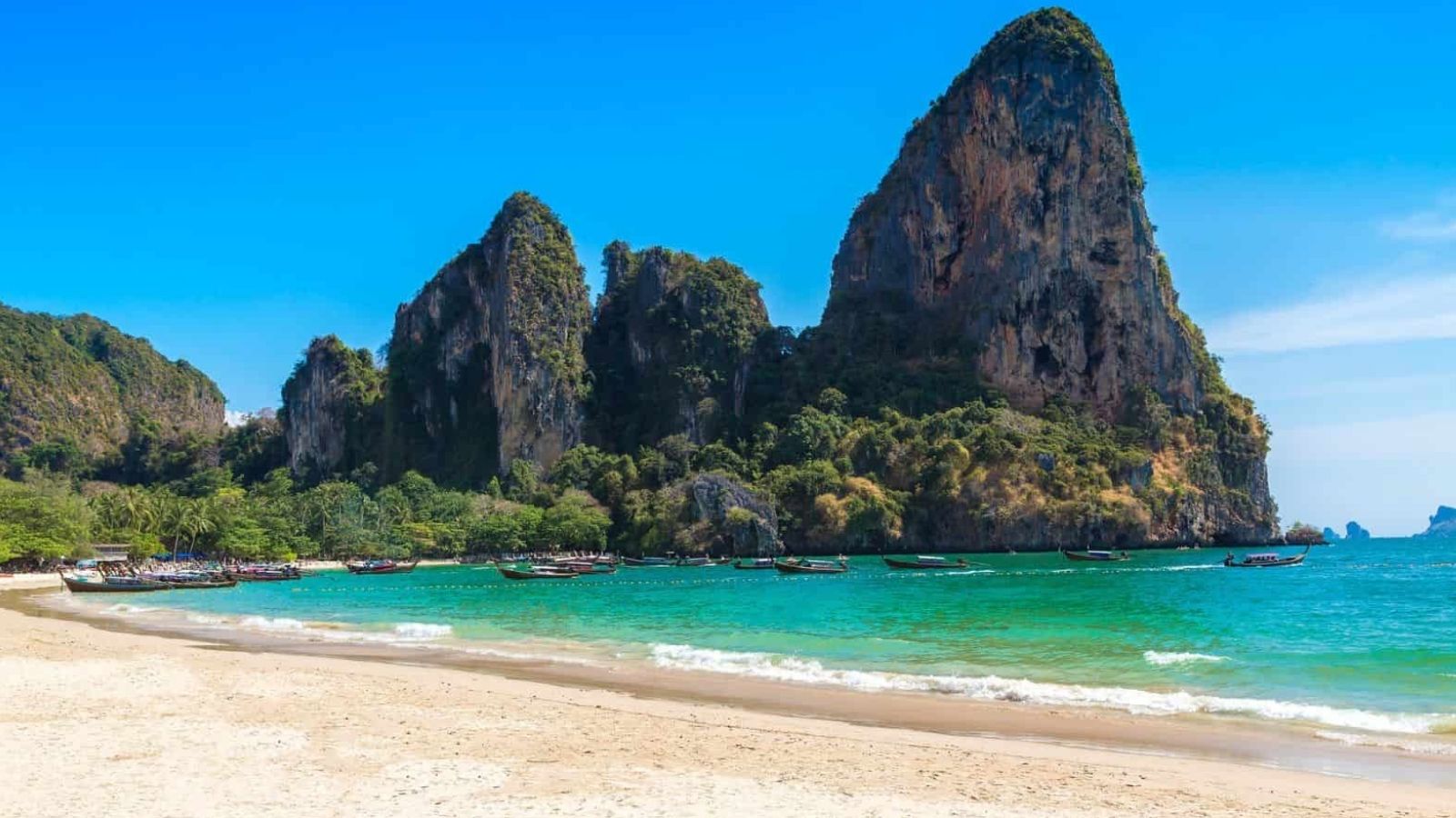 Sandy beach with turquoise water, traditional boats, and limestone cliffs beneath a clear blue sky.